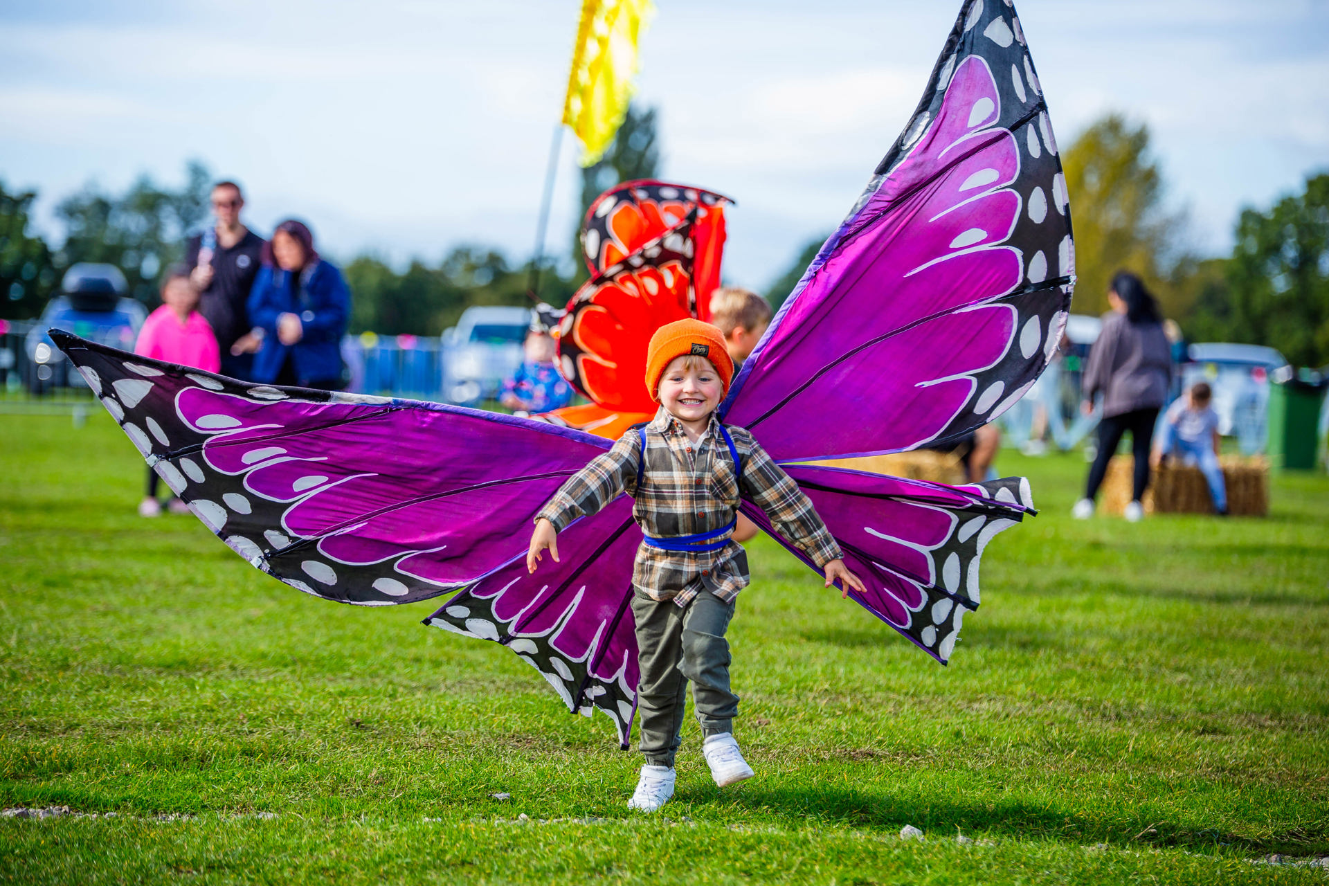 Photo shows a young boy wearing a set of pink butterfly wings. He is grinning as he runs across a field